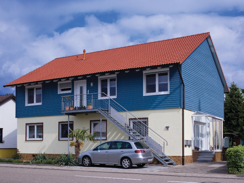 Ein zweistöckiges Haus mit blauem Obergeschoss, gelbem Erdgeschoss und rotem Dach, vor dem ein silbernes Auto geparkt ist.